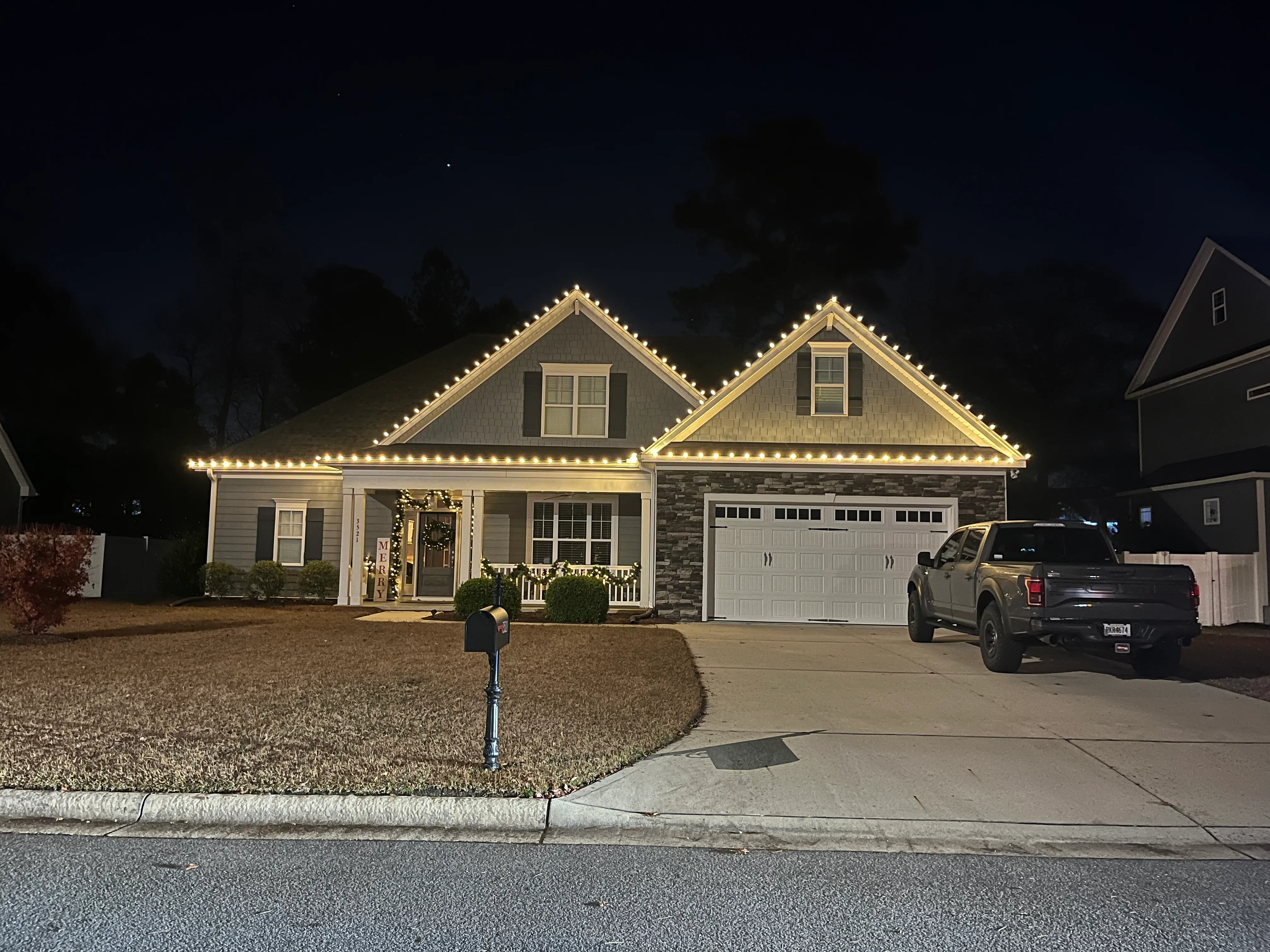 A gray house at night decorated with warm white Christmas lights outlining the roof. The porch is adorned with garlands and a wreath on the door, with a vertical sign reading 'MERRY'. A black mailbox stands on the lawn, and a dark pickup truck is parked in the driveway.