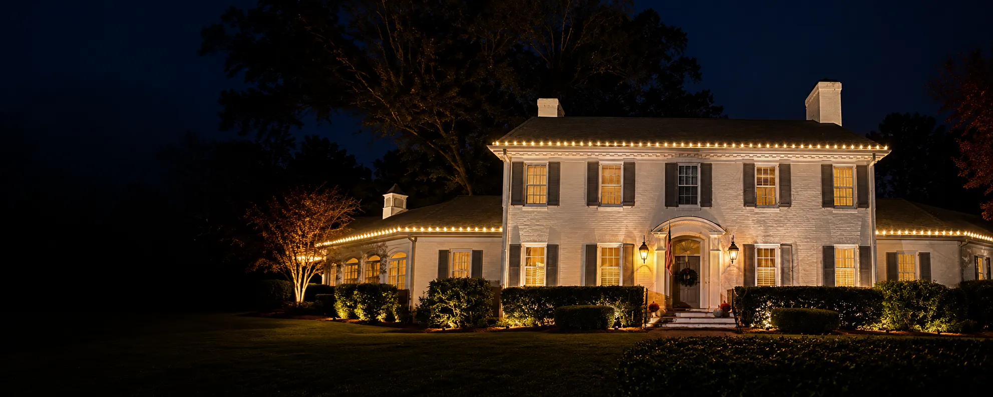 Washington, NC house lit at night with warm lights outlining the roof and illuminating bushes and trees in front, windows glowing with indoor light, and a wreath on the front door installed by Walston Evans Home Services
