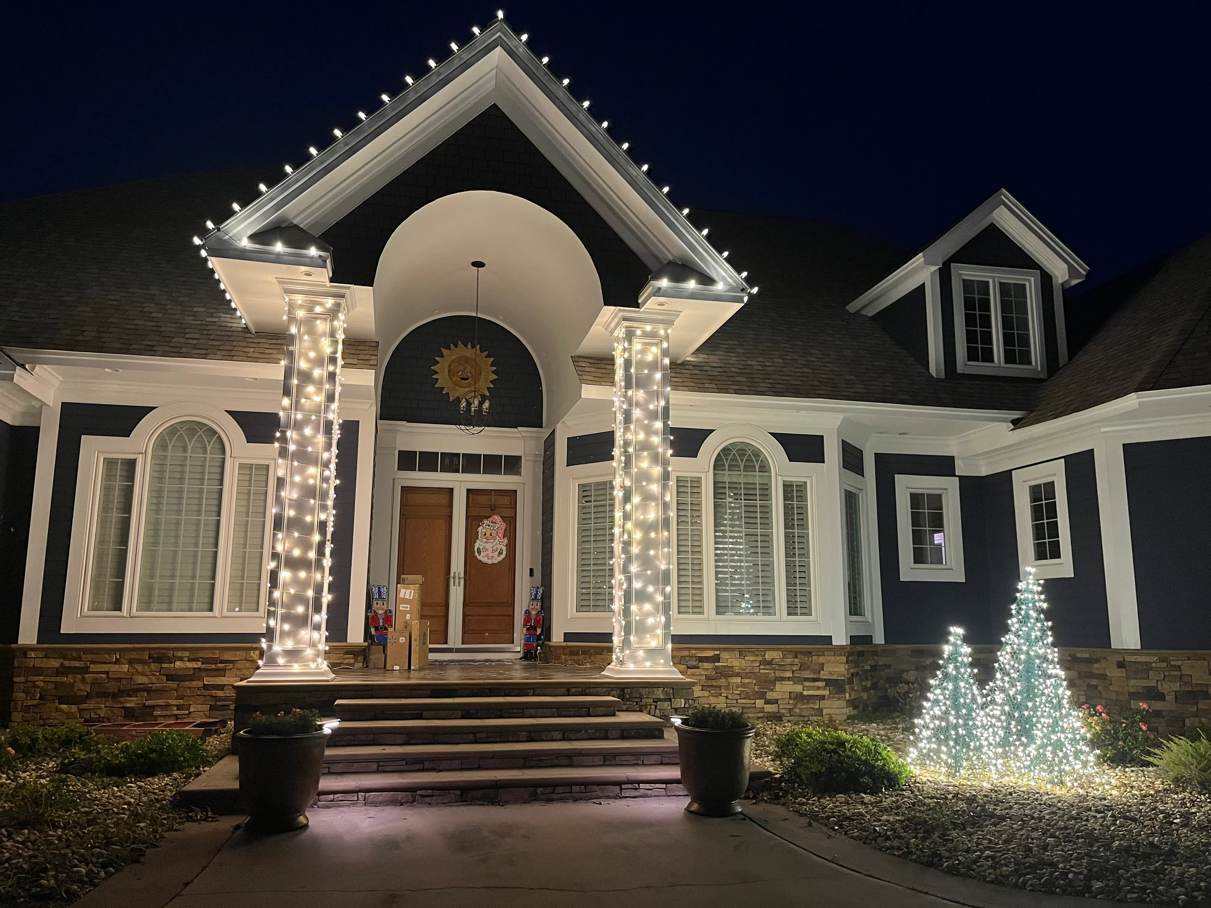 Night view of a Morehead City, NC house entrance with two white pillars wrapped in string lights, a double wooden front door decorated with a Santa ornament, two toy soldier decorations, and three small Christmas trees lit with white lights on the right side garden installed by Walston Evans Home Services