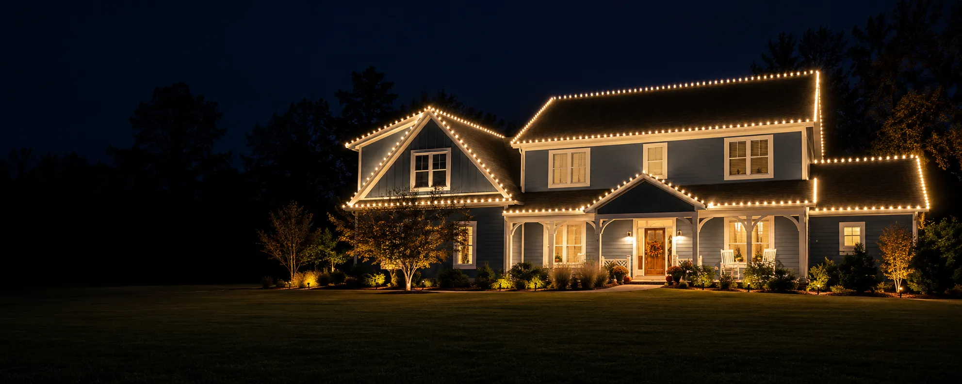 Large blue farmhouse in Morehead City, NC lit with warm string lights outlining the roof and porch at night, surrounded by illuminated bushes and trees with Christmas lights installed by Walston Evans Home Services.