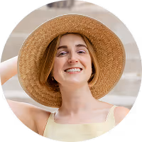 A portrait of Allison Press, a white woman with a reddish blonde bob. She's wearing a straw sun hat, yellow tank top. She holds the hat on with one hand at the back of her head as she leans back and smiles at the camera.
