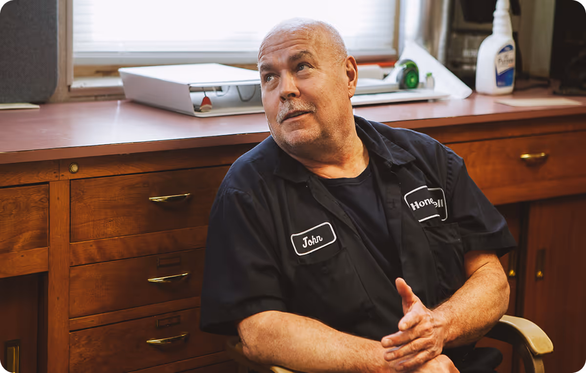 An older white man with a short white mustache and buzzed white hair talks to someone off camera. He's wearing a black, button down trades uniform that says "John" and "Honeywell." He's in a simple office, sitting in front of a desk.
