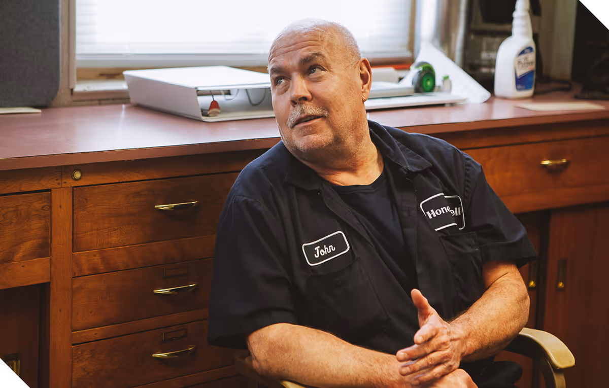 An older white man with a short white mustache and buzzed white hair talks to someone off camera. He's wearing a black, button down trades uniform that says "John" and "Honeywell." He's in a simple office, sitting in front of a desk.
