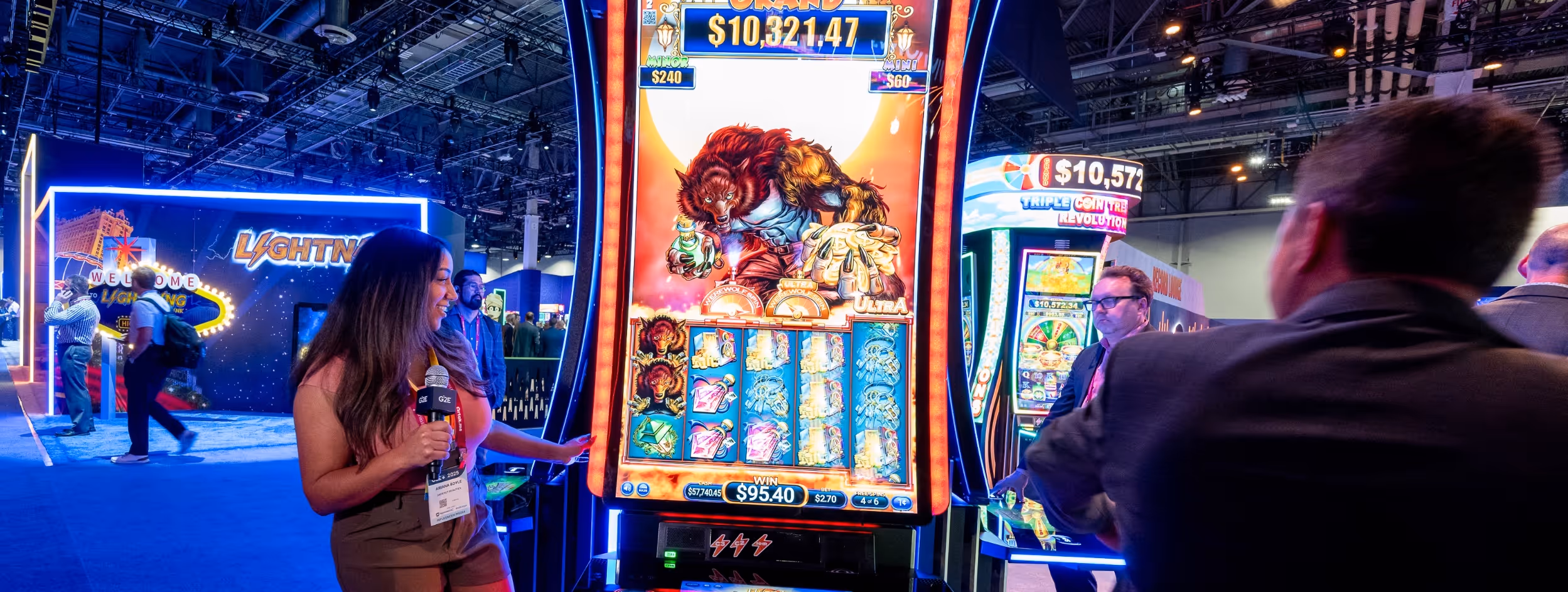 A woman holding a microphone stands next to a brightly lit slot machine featuring a werewolf design at a gaming convention.