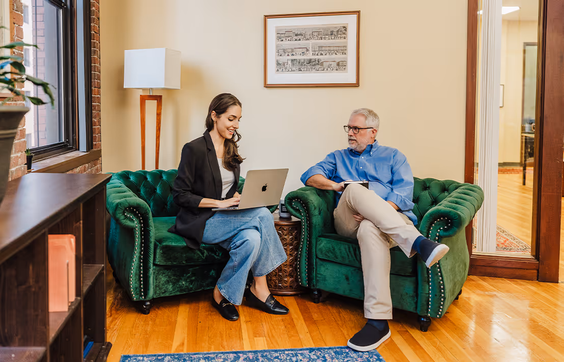 Two MHJP team members sitting on green couches while looking at a laptop.