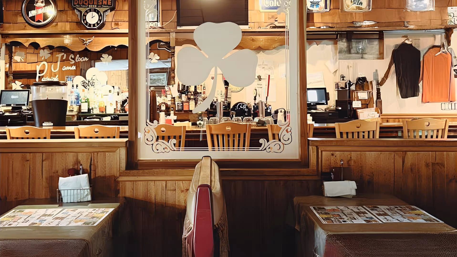Cozy tavern interior with wooden booths and tables, adorned with menus. A glass panel features a clover design, revealing a bar with bottles and decor. Warm lighting creates an inviting atmosphere.