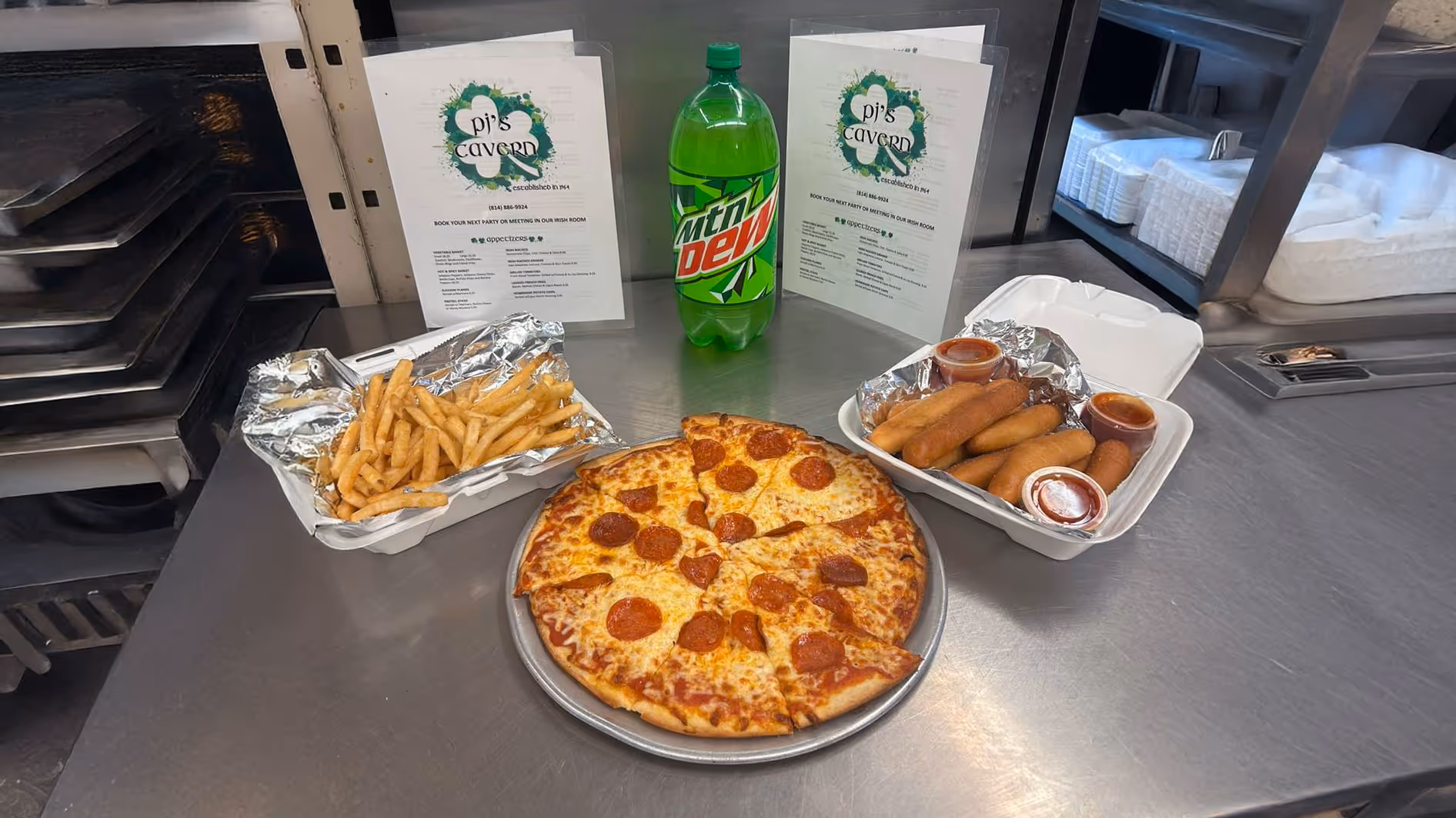 A pepperoni pizza on a metal tray is flanked by fries and mozzarella sticks in takeout boxes. A Mountain Dew bottle and menus are in the background.