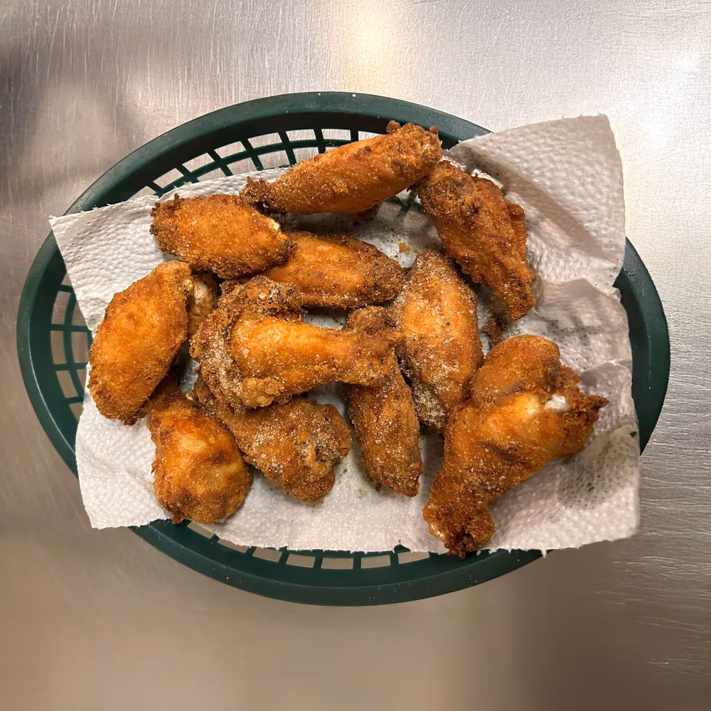 A basket of golden-brown, crispy fried chicken wings on paper towels, resting on a shiny metal surface. The wings appear crunchy and appetizing.