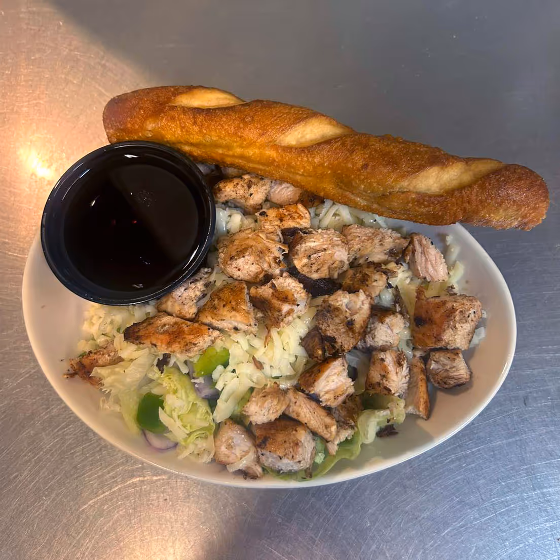 A white bowl, on a metal surface, filled with grilled chicken pieces and shredded lettuce, topped with shredded cheese. A side of dark sauce is in a cup, with a bread stick resting on top.