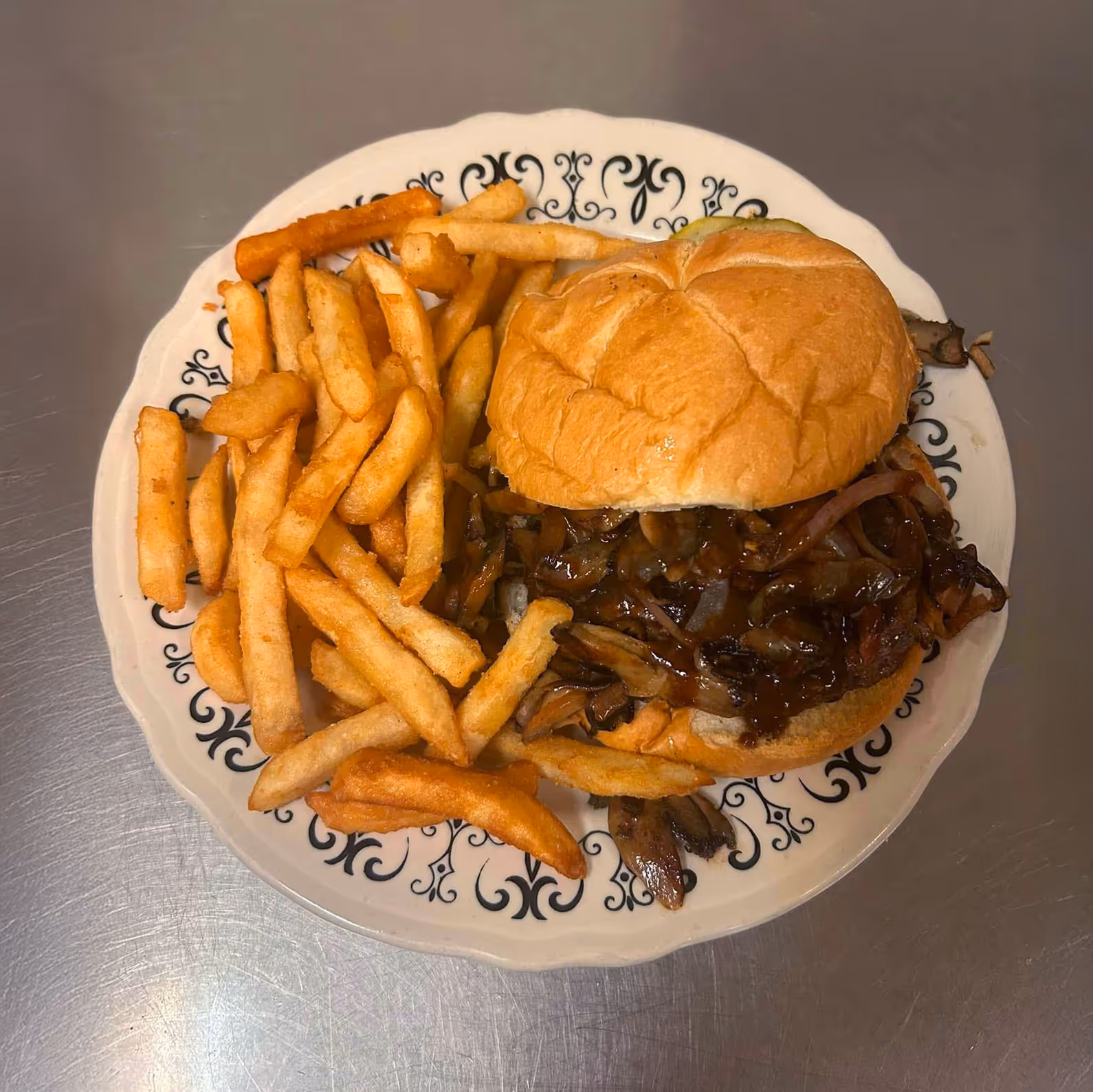 A juicy burger with caramelized onions on a bun, served with golden, crispy fries on a plate, set against a stainless steel surface.