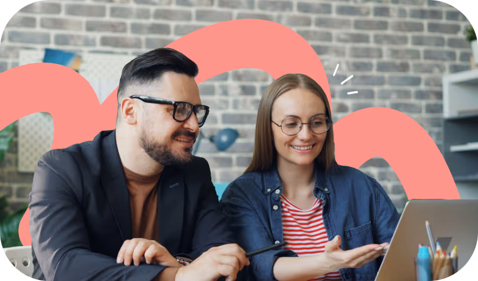 Two people with glasses smiling and discussing while looking at a laptop in a workspace with a brick wall background.