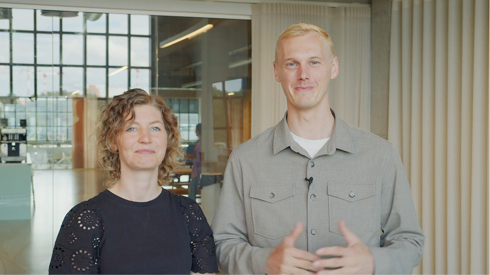 A man and a woman standing indoors with a glass wall and curtains behind them.