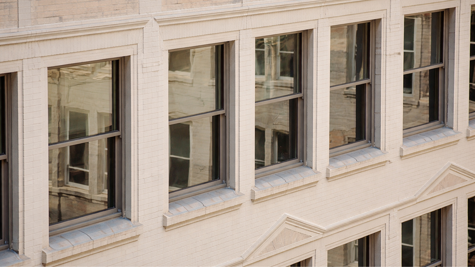 Close-up of a beige brick building facade with multiple dark-framed windows reflecting another building.