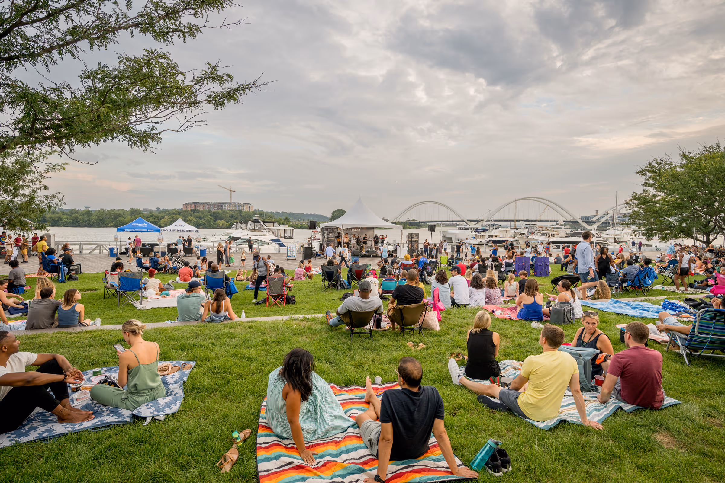 A crowd on blankets and lawn chairs at a Friday night concert on the Yards Park lawn, with the river and a bridge behind
