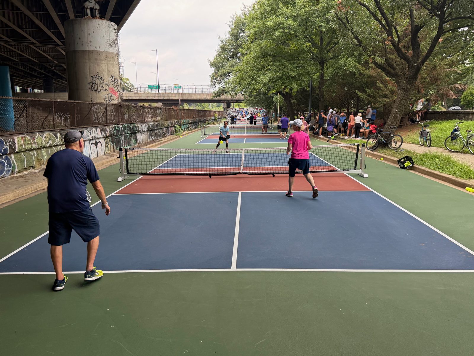 Doubles players on the blue and green courts, with more courts and the freeway overpass beyond