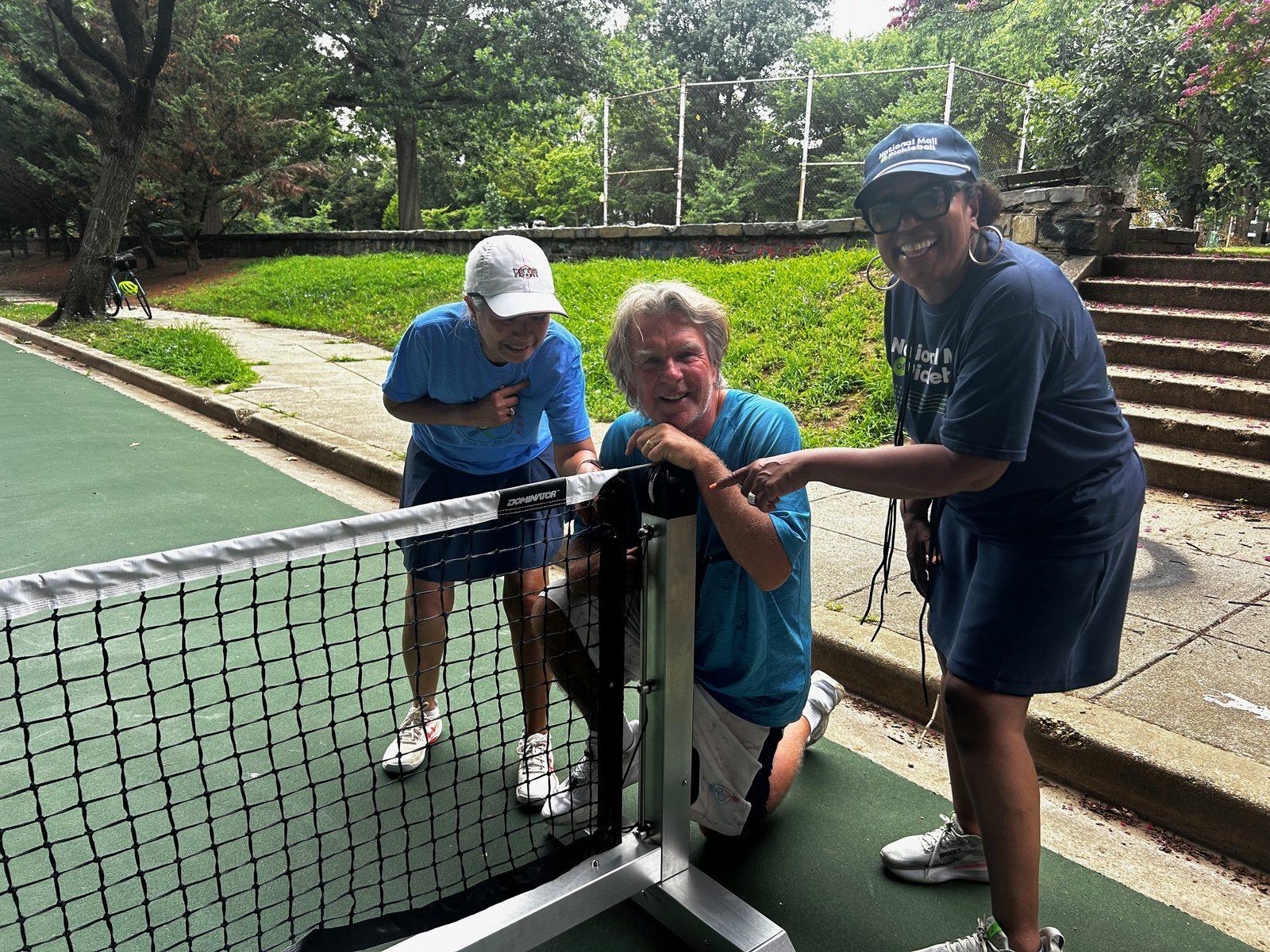 Three volunteers smiling as they assemble a net on the new courts
