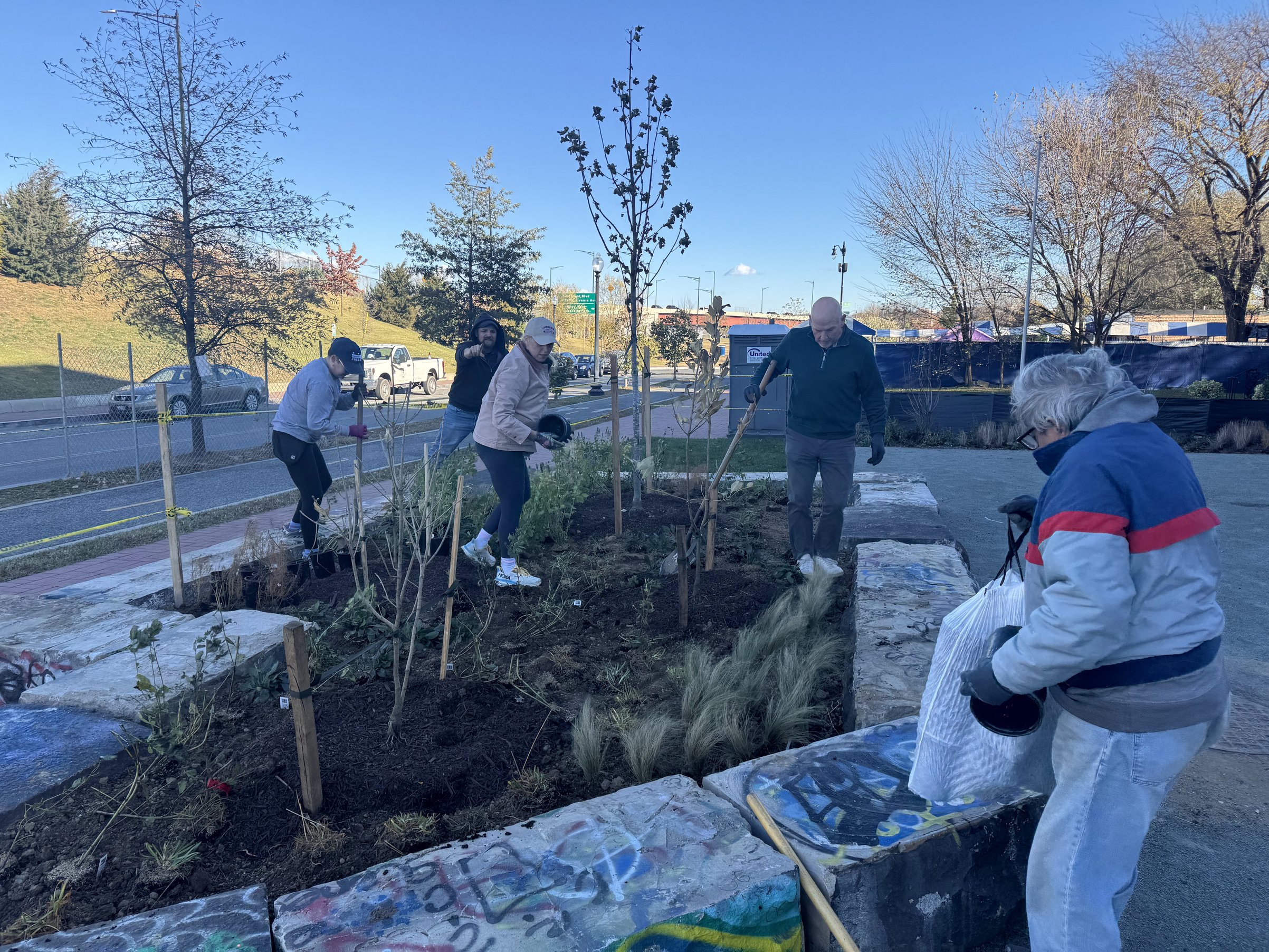 Neighbors plant young trees and perennials inside the portal-stone beds at Emblem Plaza's community planting day, November 16, 2025