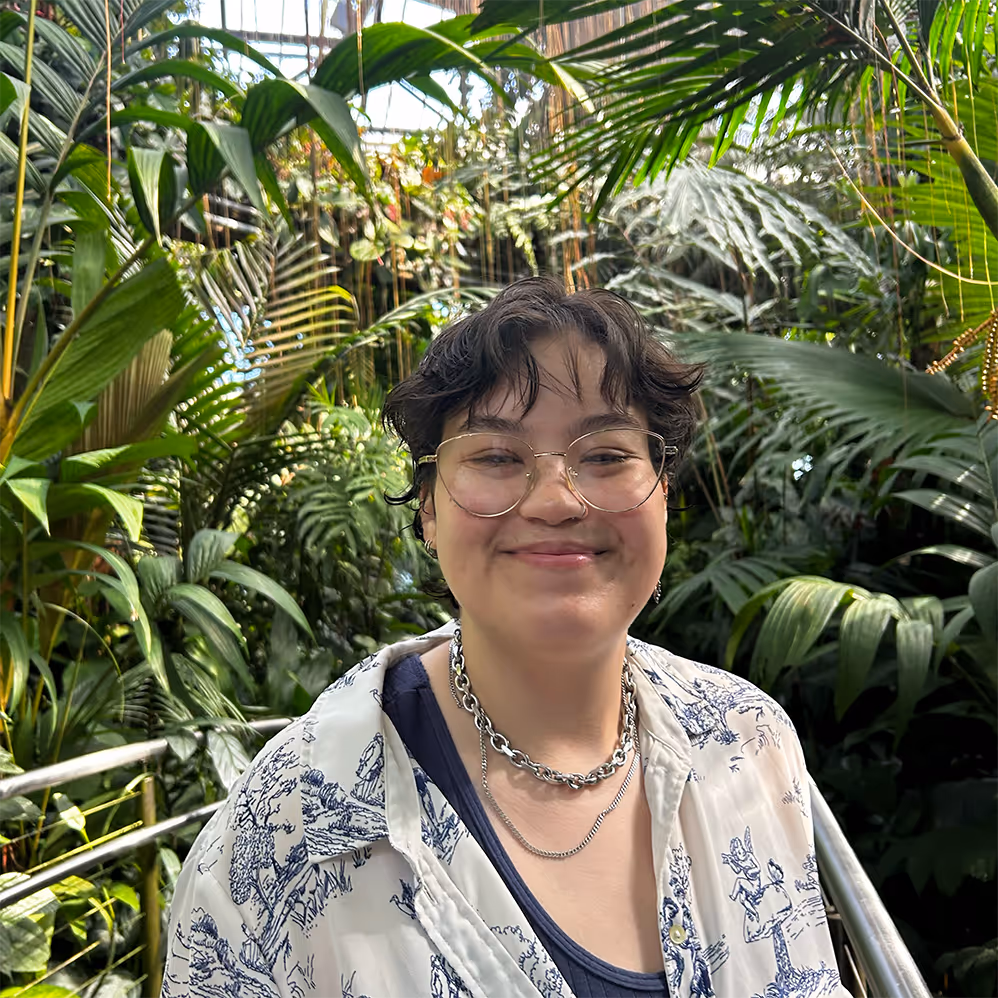 Suhayla with short dark hair and glasses smiling, standing on a walkway surrounded by lush green tropical plants.