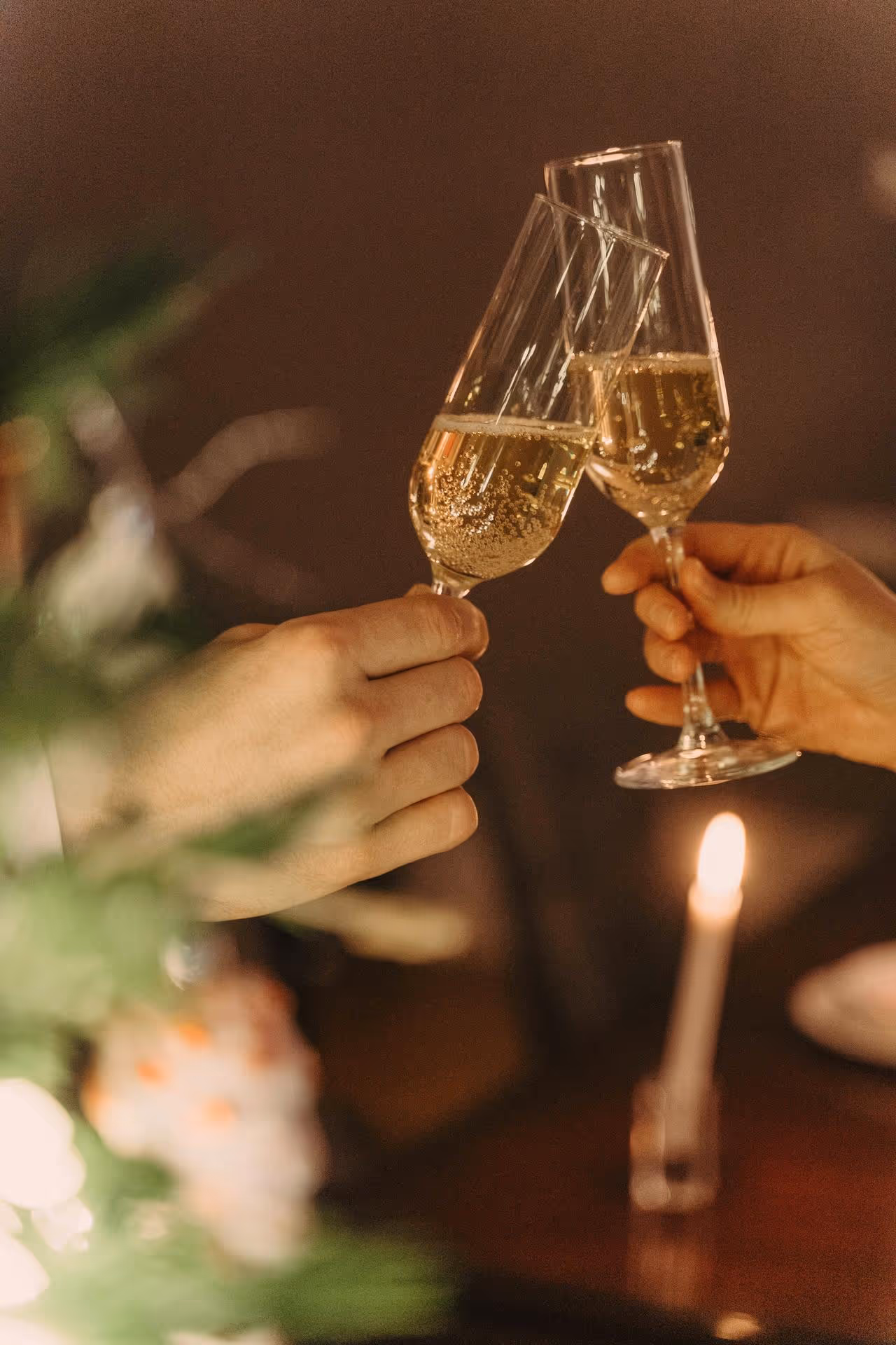Two hands clink champagne glasses in a warm, dimly lit setting. A candle flickers in the background, adding a festive, celebratory mood.
