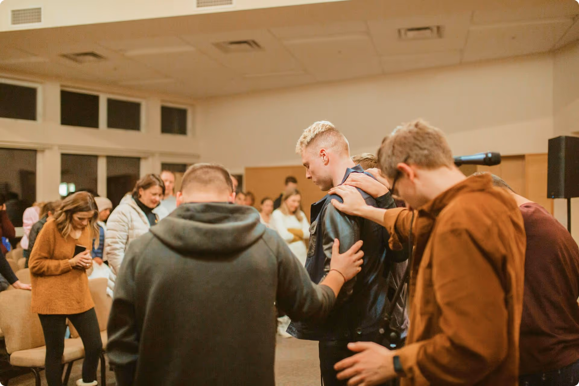 A group of people stands in a room, with several placing hands on one person's shoulders in a supportive gesture. The mood is calm and focused.