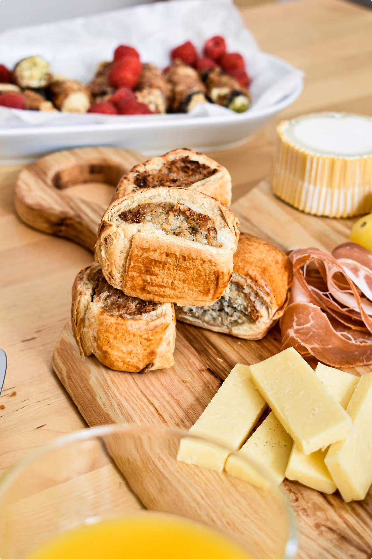 A wooden board with sausage rolls, sliced cheese, and cured meat on a table. In the background, a tray of raspberries and other snacks. Cozy brunch setting.