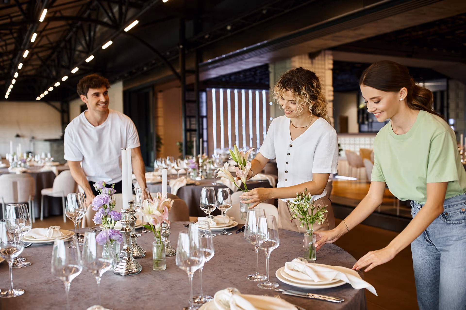 Three people set a table for an elegant event, arranging flowers and napkins. The atmosphere is focused and collaborative in a well-lit venue.