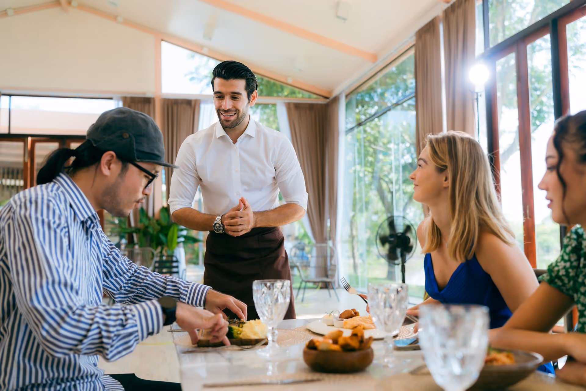 A smiling waiter in a white shirt stands by a table with three people dining in a bright, elegant restaurant. The atmosphere is warm and friendly, with large windows showing greenery outside.