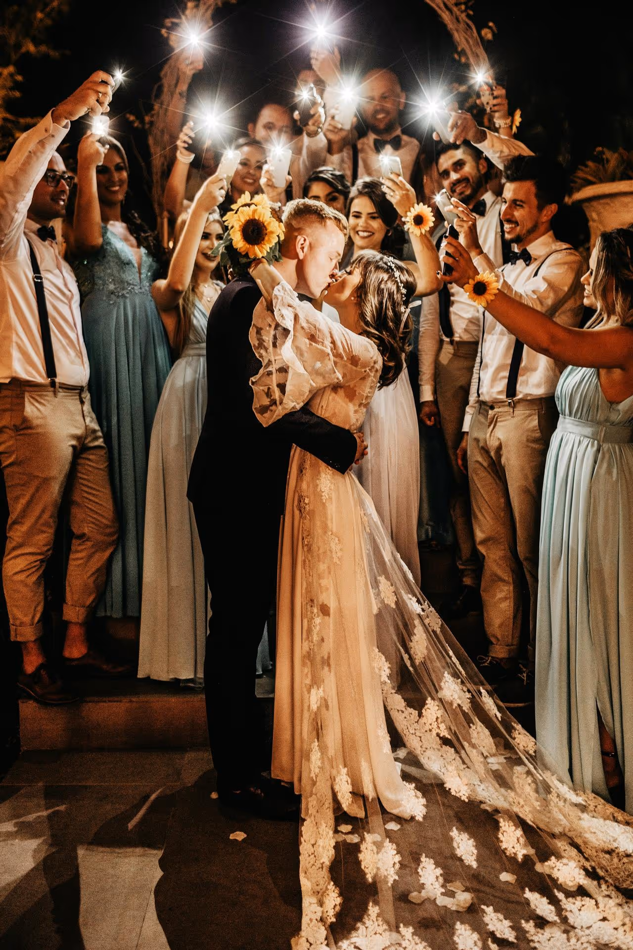 Bride and groom share a kiss under an arch, surrounded by joyful guests holding sunflowers and sparklers. The scene is warm and celebratory.