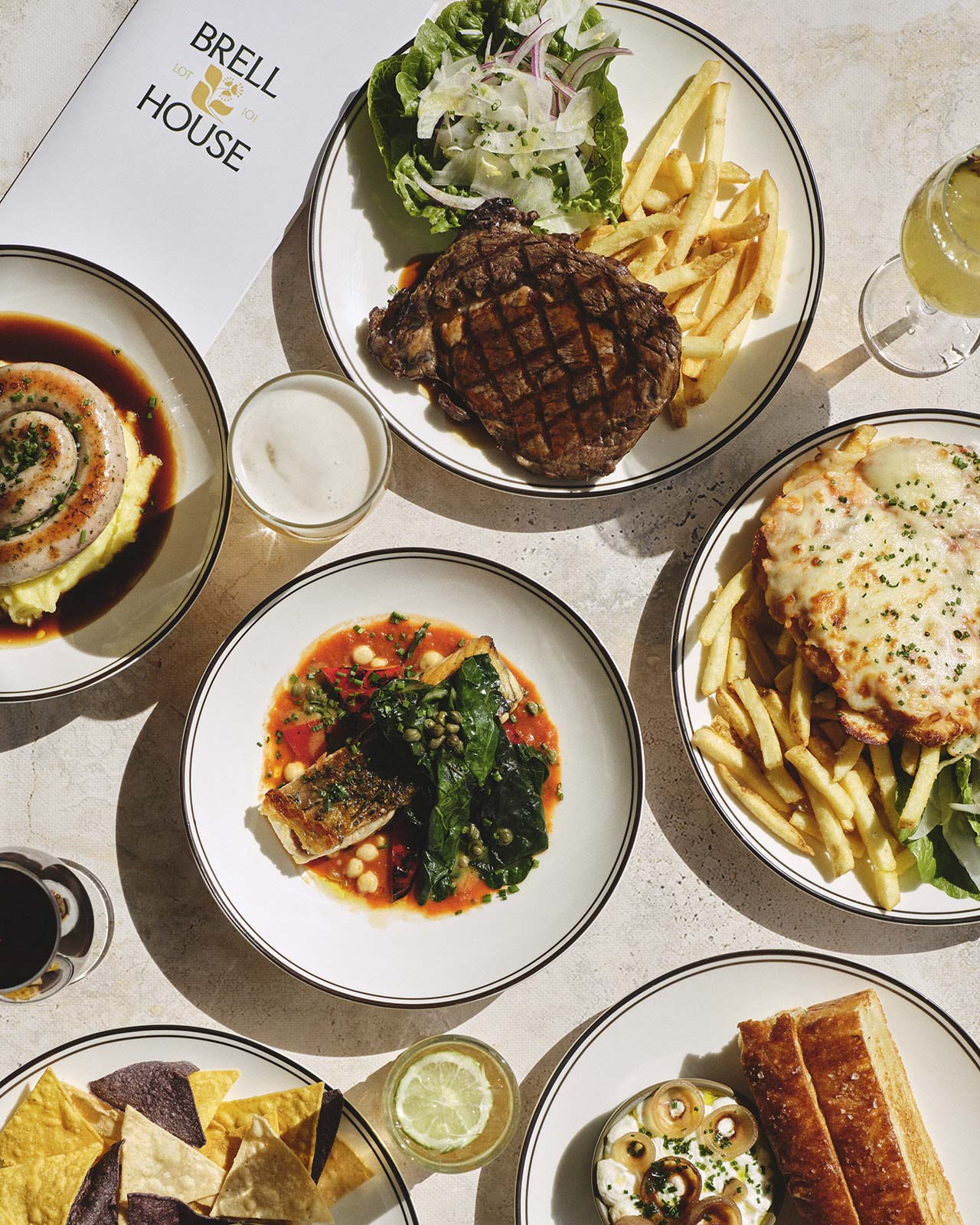 A marble table with various appetizers, including stuffed rolls, bruschetta, and a vibrant dish in red sauce. Five hands reach for plates, creating a social, lively atmosphere. Drinks include cocktails and water.