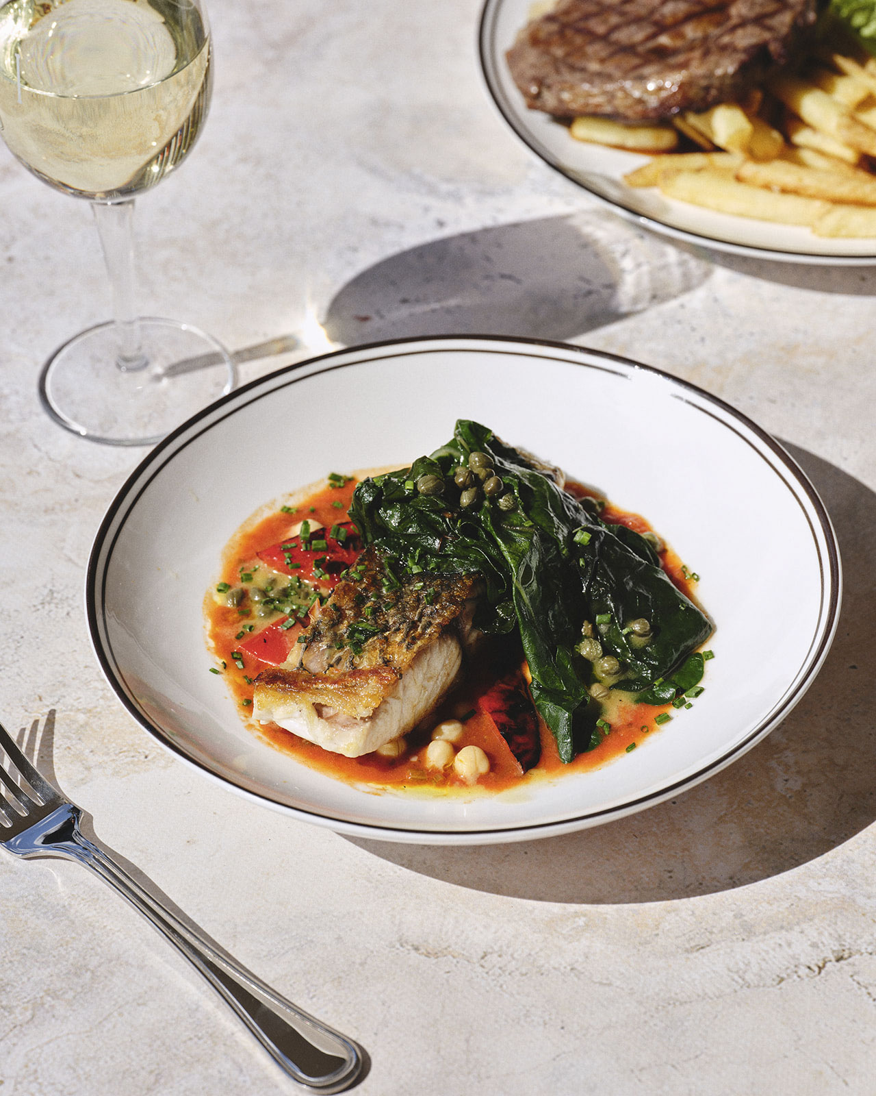 Two beautifully plated dishes on textured plates. One features roasted meat and greens, the other has colorful root vegetables and nuts. Elegant and appetizing.