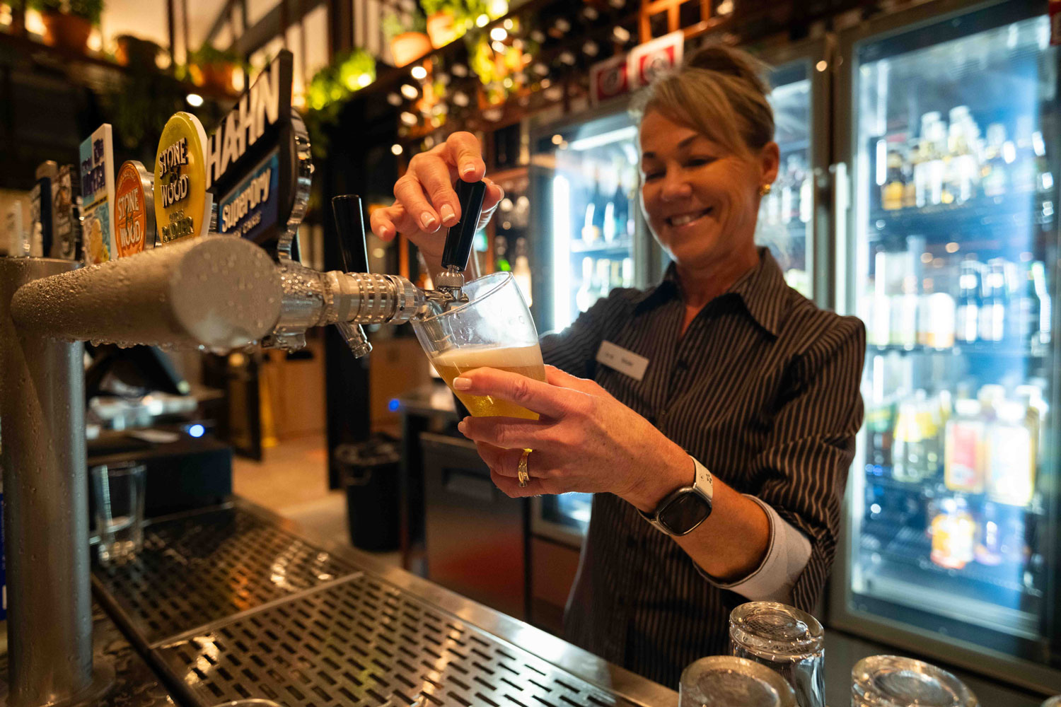 A smiling waiter in a white shirt stands by a table with three people dining in a bright, elegant restaurant. The atmosphere is warm and friendly, with large windows showing greenery outside.