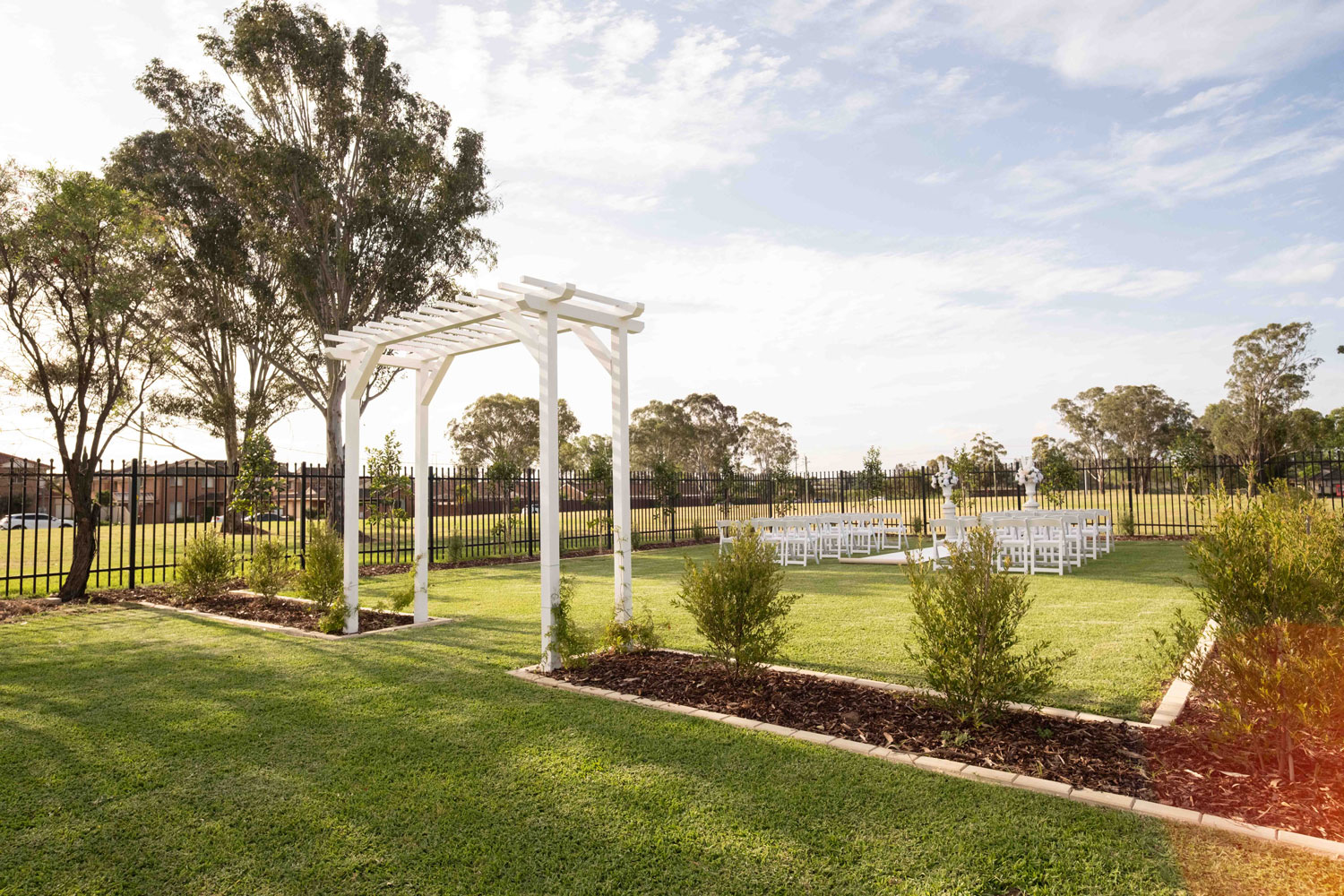 A bride in a flowing white gown, holding a bouquet, walks down a cobblestone path through a lush garden toward a gathering of seated guests.