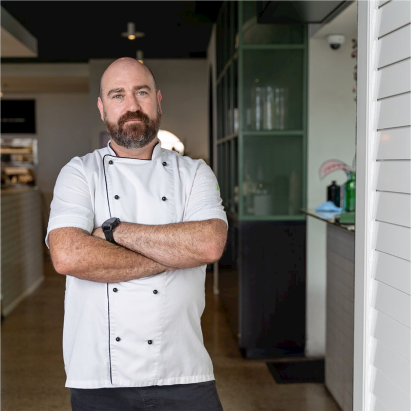 Chef focused on plating food in a dimly lit kitchen. Wearing a white uniform and apron, the chef uses a pot, conveying concentration and dedication.