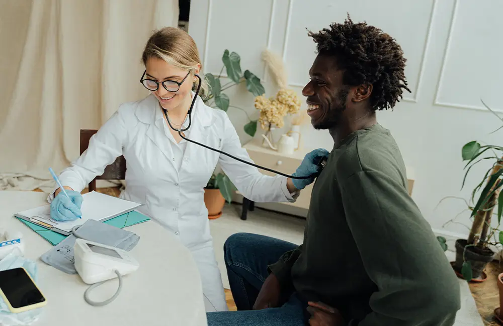 A female doctor using a stethoscope to listen to the back of a smiling male patient seated in a medical office.