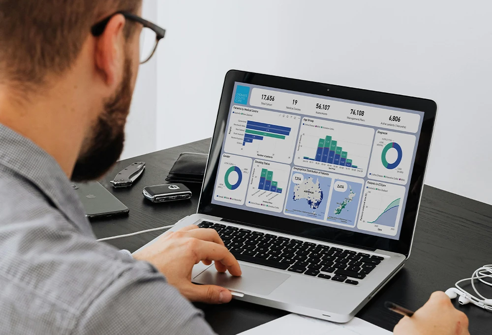 Man with glasses working on a laptop displaying medical center statistics and data visualizations.
