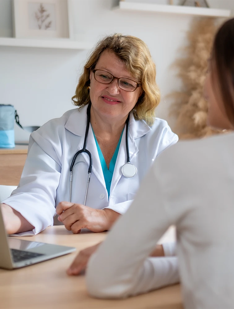 Female doctor with stethoscope consulting a patient across a desk with a laptop.
