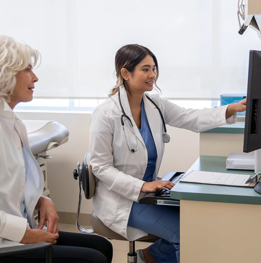Female doctor in white coat with stethoscope pointing at computer screen while consulting with older female patient in a medical office.