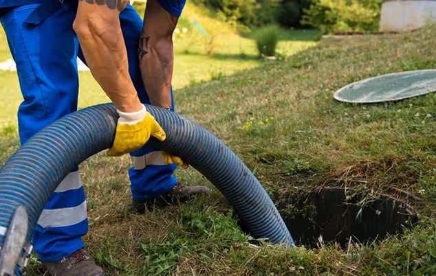 A man in blue overalls cleans a drain using a hose, focused on removing debris and ensuring proper drainage.
