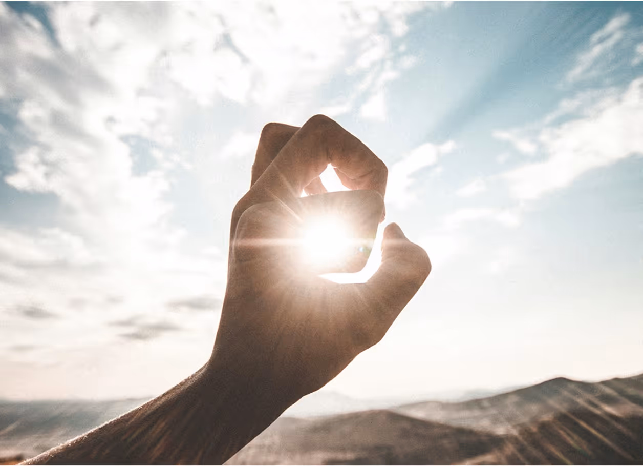 Hand forming an OK gesture with sunlight shining through the circle against a blue sky with clouds.