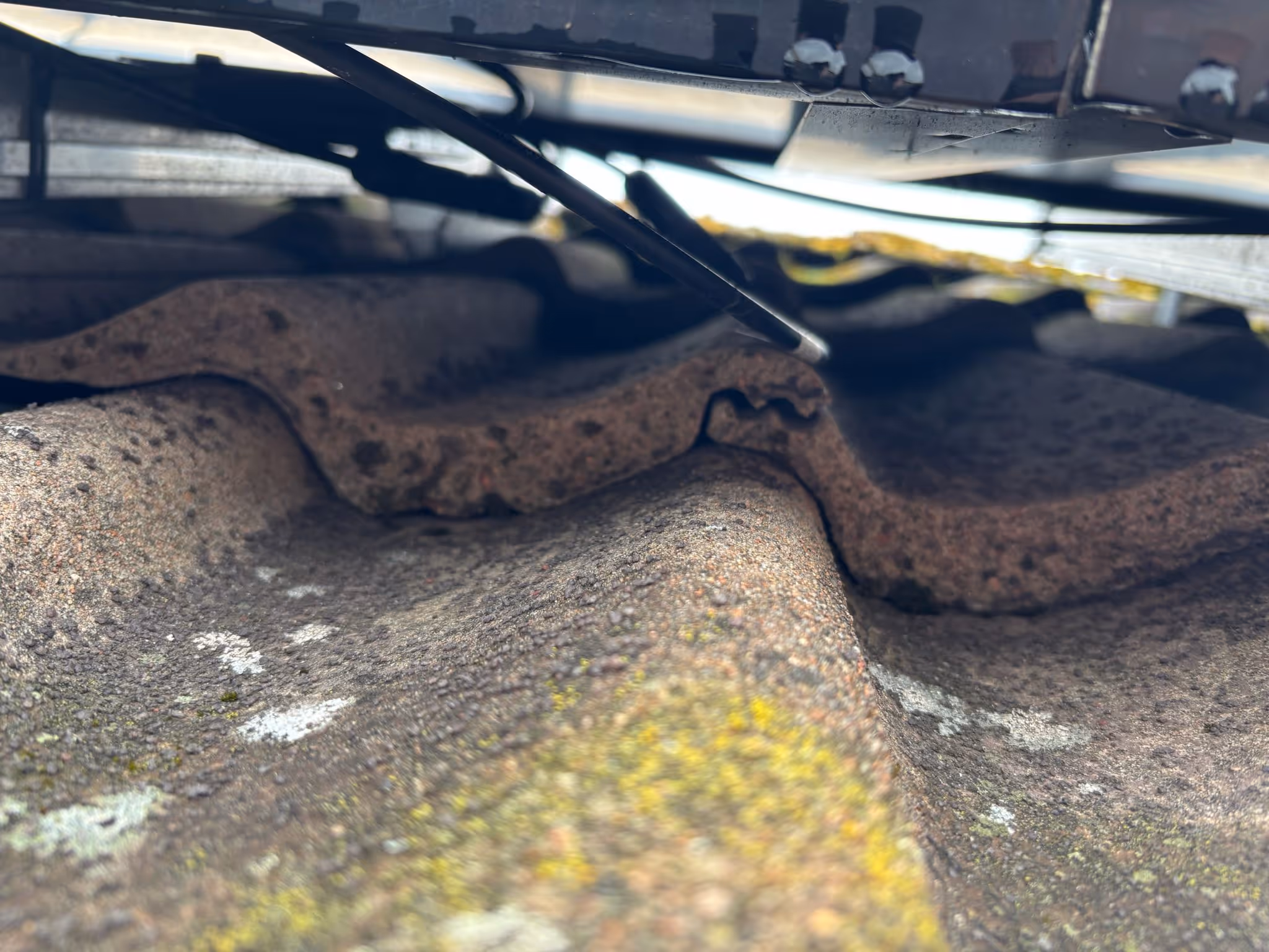 Närbild av mossig och väderbiten vågformad takpanna under en takkant med hängande lösa kablar.