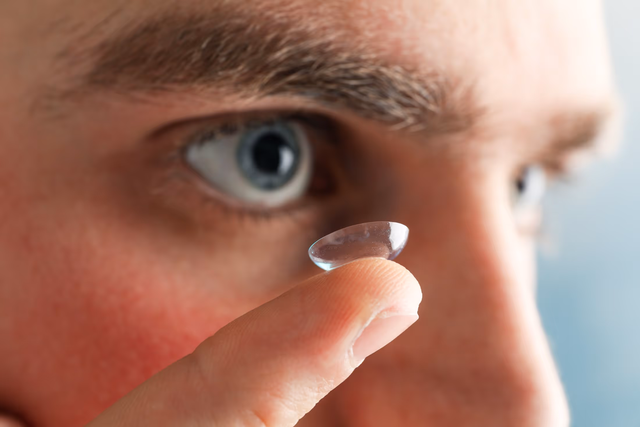 Close-up of a contact lens on a fingertip near a blue eye