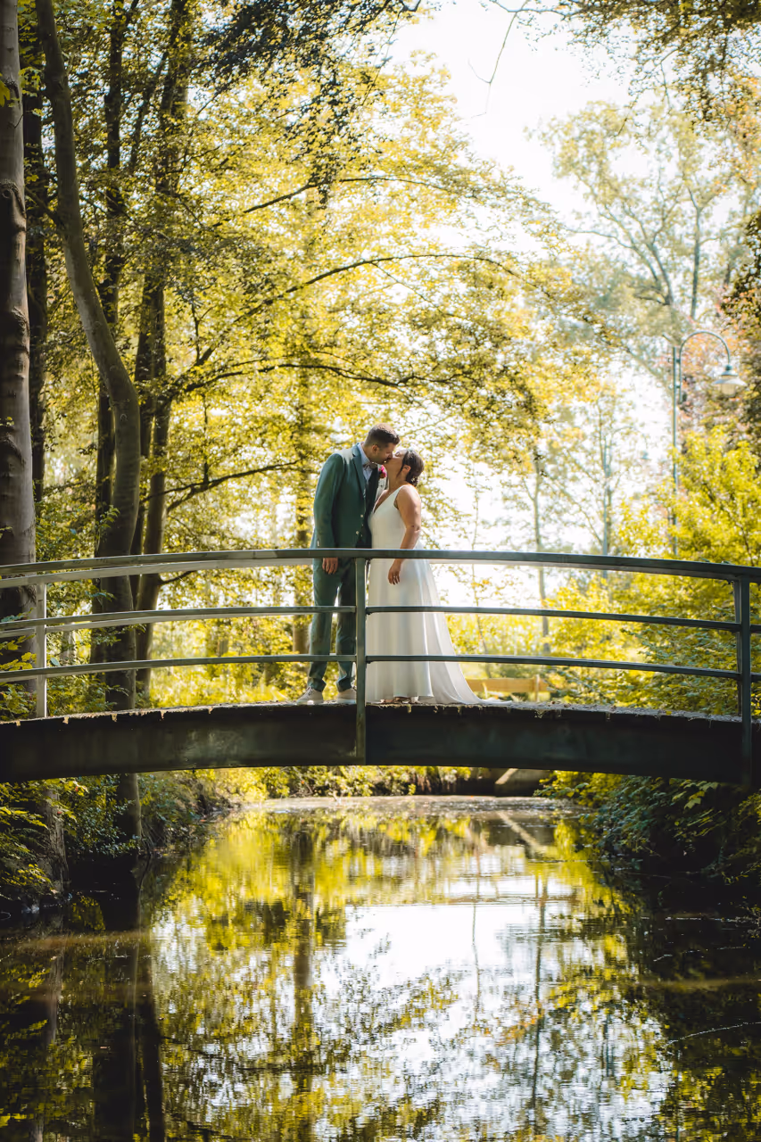 Witte bruid en bruidegom in groen kostuum kussen elkaar op een brug in een park.