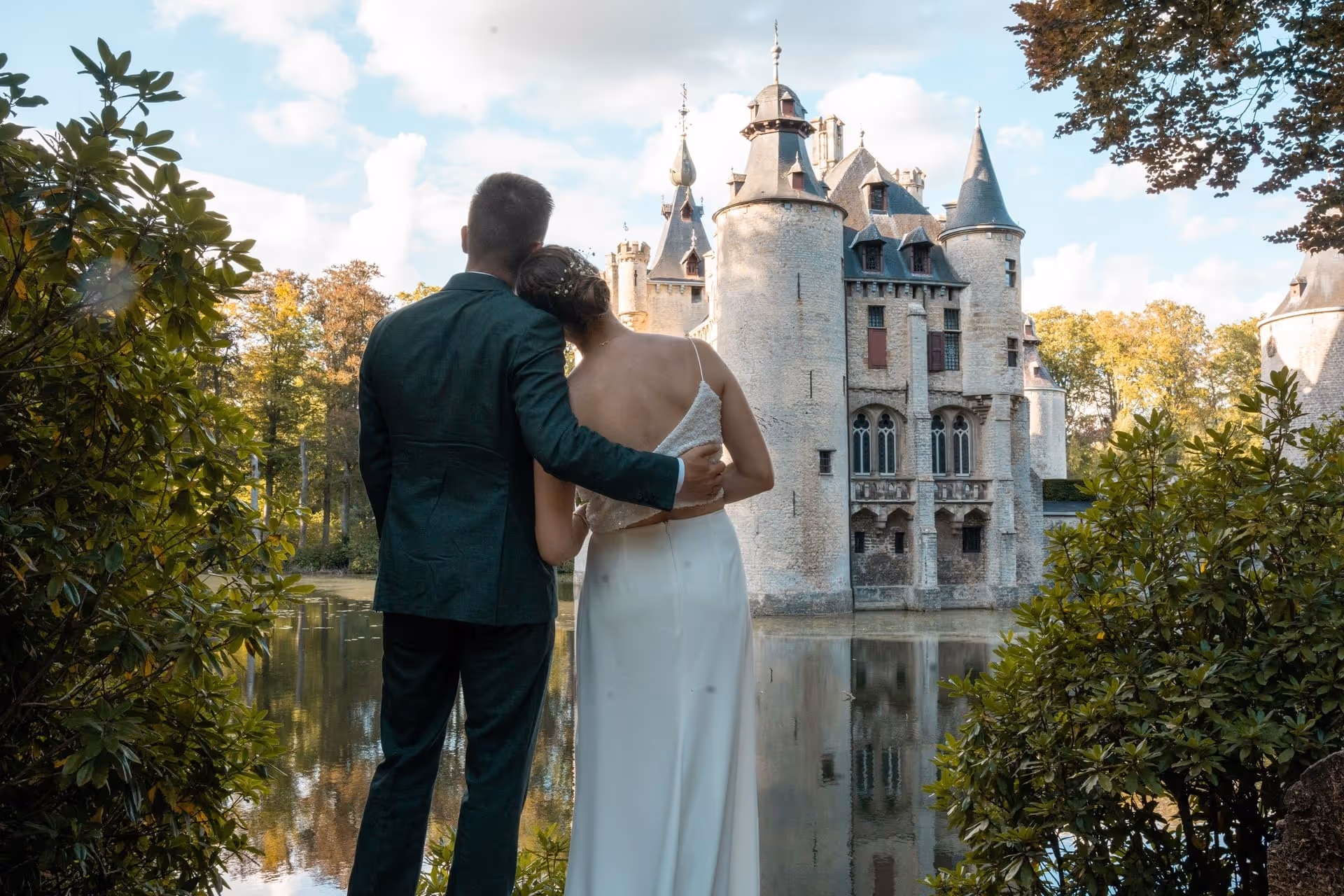Koppel in trouwkledij omhelst elkaar en kijkt naar een historisch stenen kasteel omgeven door een gracht.