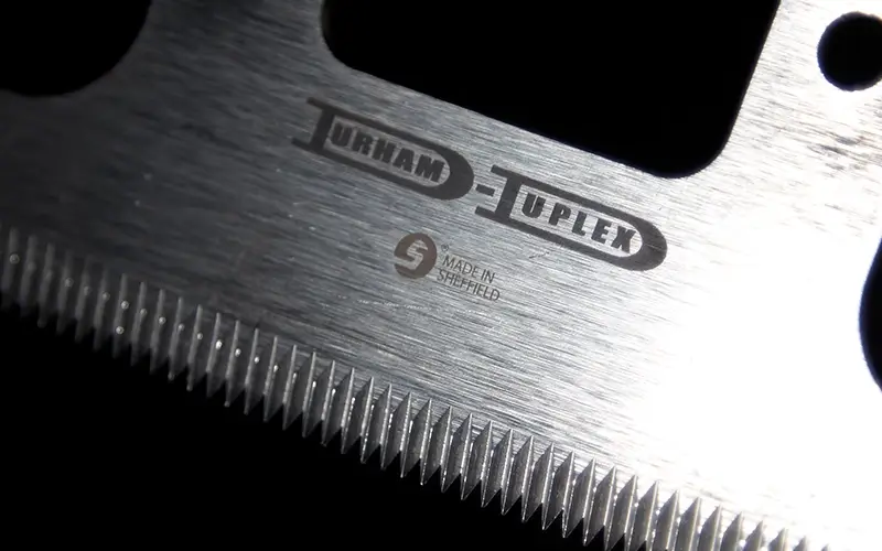 Close-up pf a serrated stainless steel blade engraved with the Durham Duplex logo, used as a precision packaging blade.