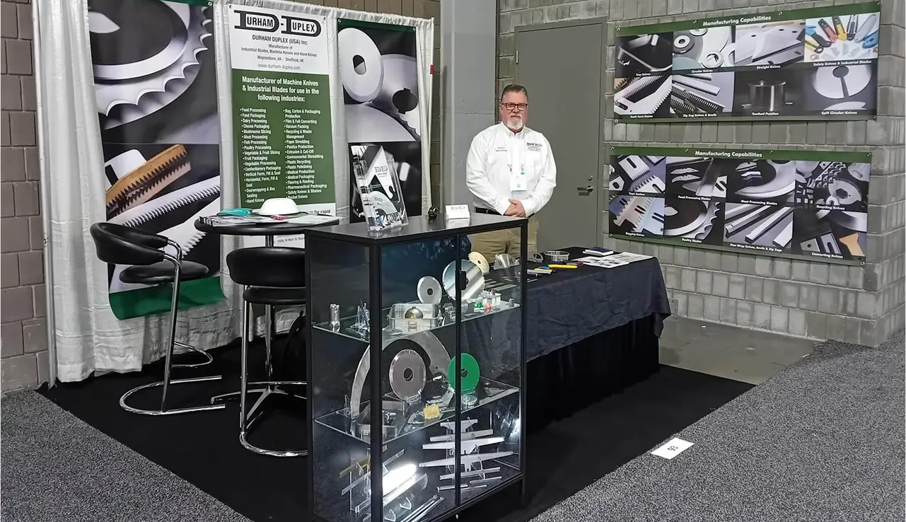 Convention stand showcasing a display of industrial knives and machine blades, with company representative standing behind the table.