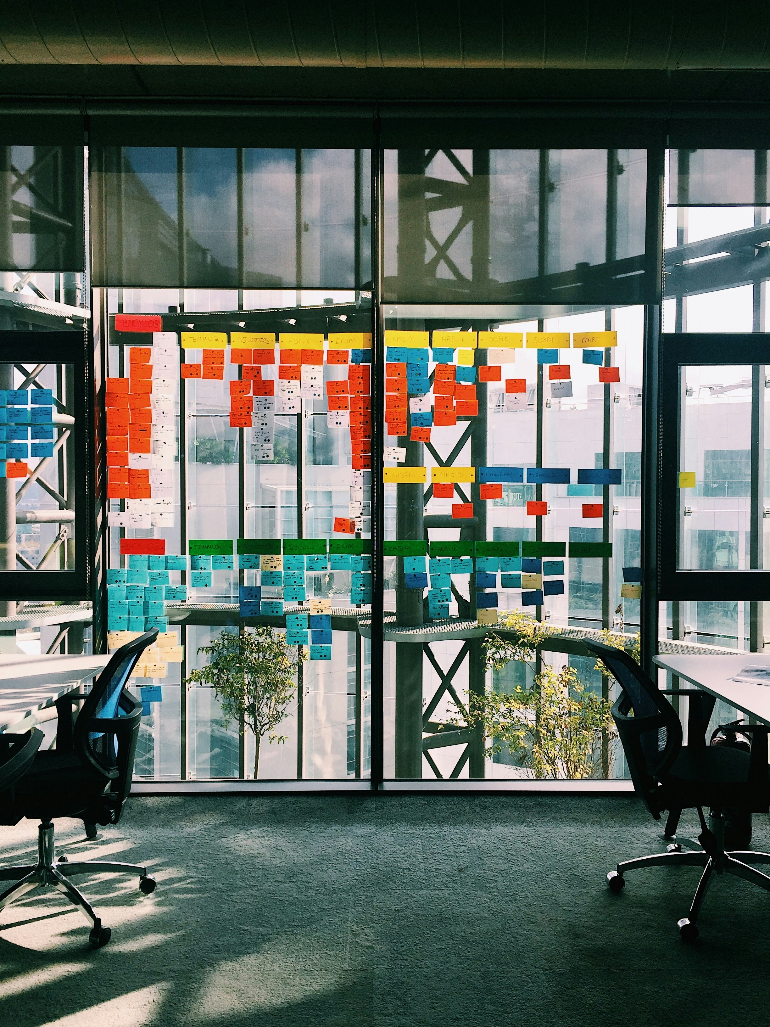 Office windows covered with colorful sticky notes for project planning with two empty office chairs nearby.