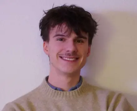 Young man with dark hair and mustache smiling, wearing beige sweater against a plain wall.