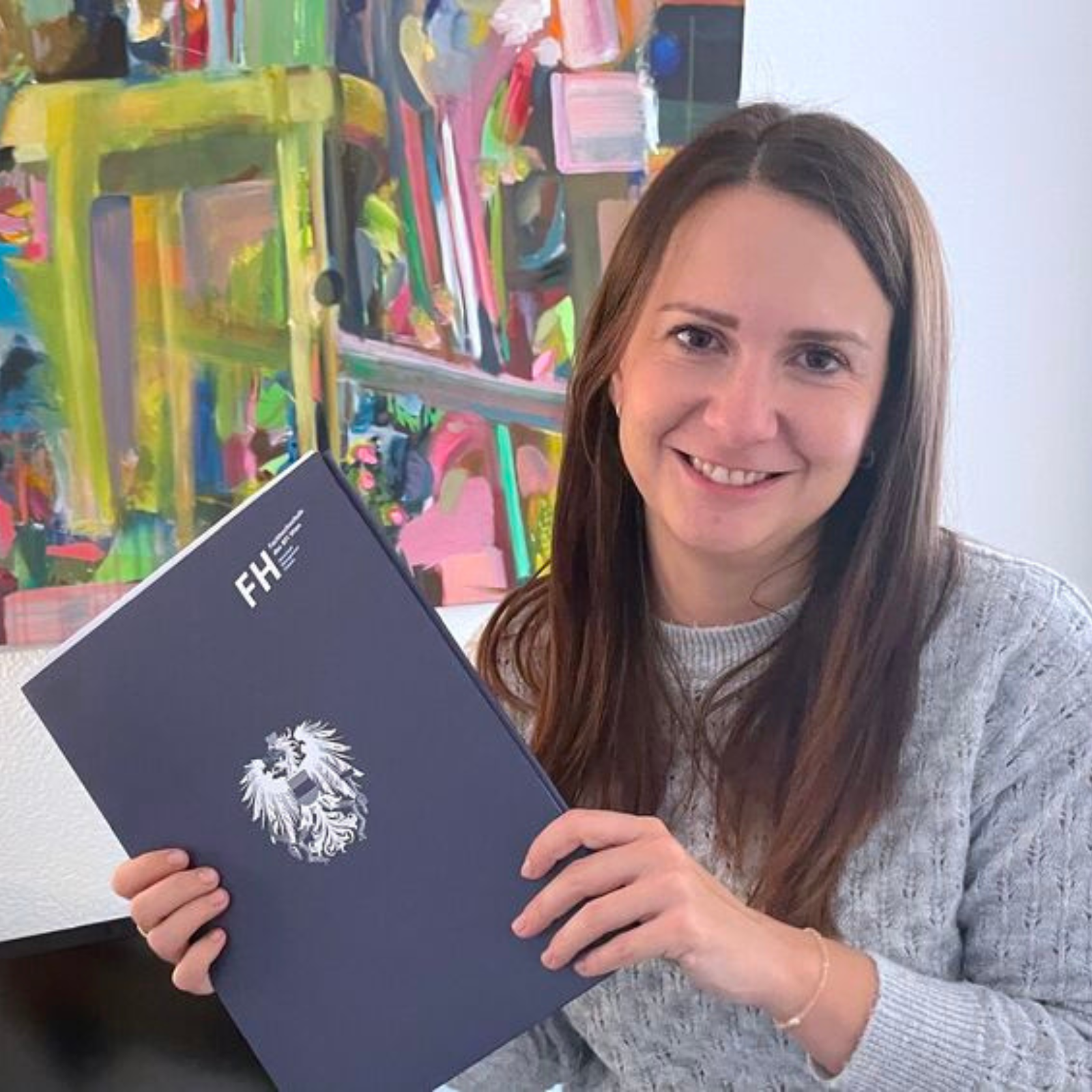 Smiling woman with long brown hair holding a navy folder with FH logo and crest in front of colorful artwork.