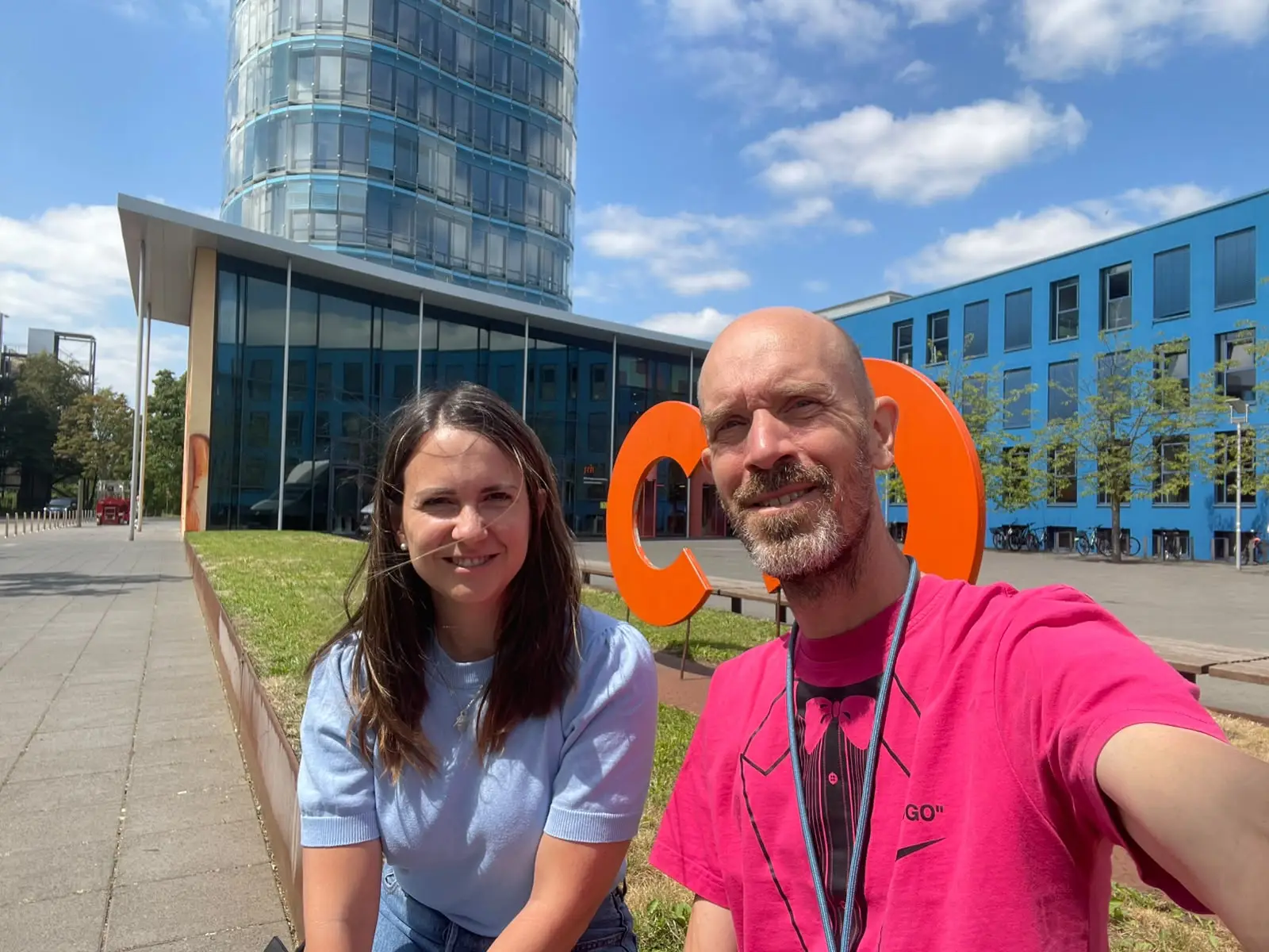 A man and a woman smiling outdoors near a modern building with glass and a large orange sculpture.