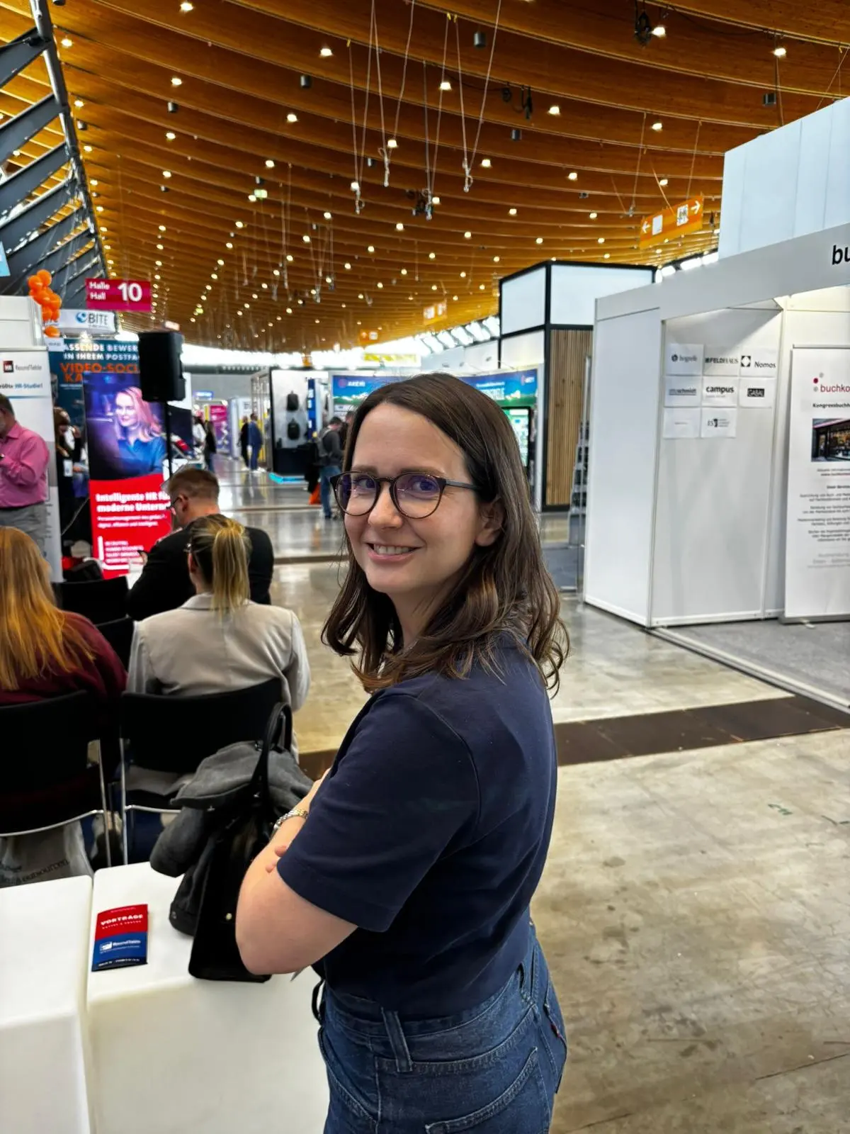 Smiling woman with glasses in a navy shirt at a busy indoor event or exhibition space.